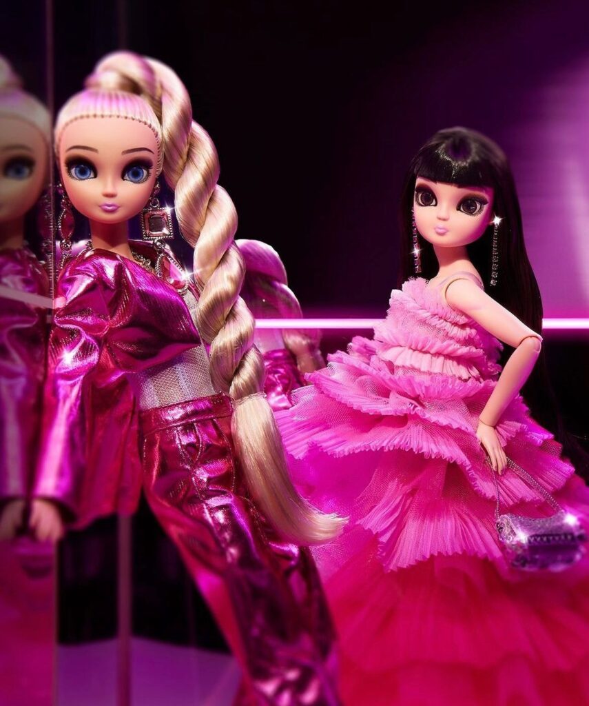 Imagem de duas bonecas barbie, uma de cabelo preto e outra de cabelo loiro, as duas estão vestidas de rosa.